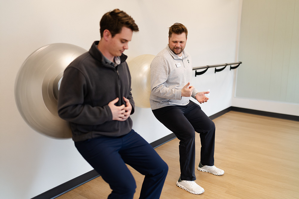 Two men are exercising indoors, each leaning against a wall with an exercise ball behind their back, performing wall squats. They appear to be interacting and smiling.