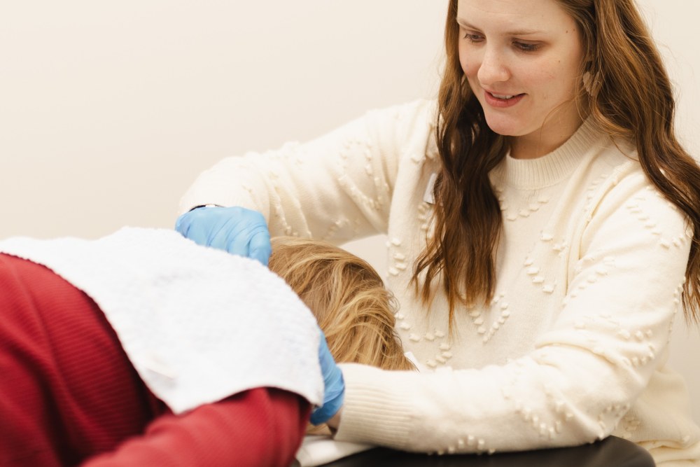 A woman in a white sweater and blue gloves examines or treats the head of a person lying face down on a table, covered with a white towel.