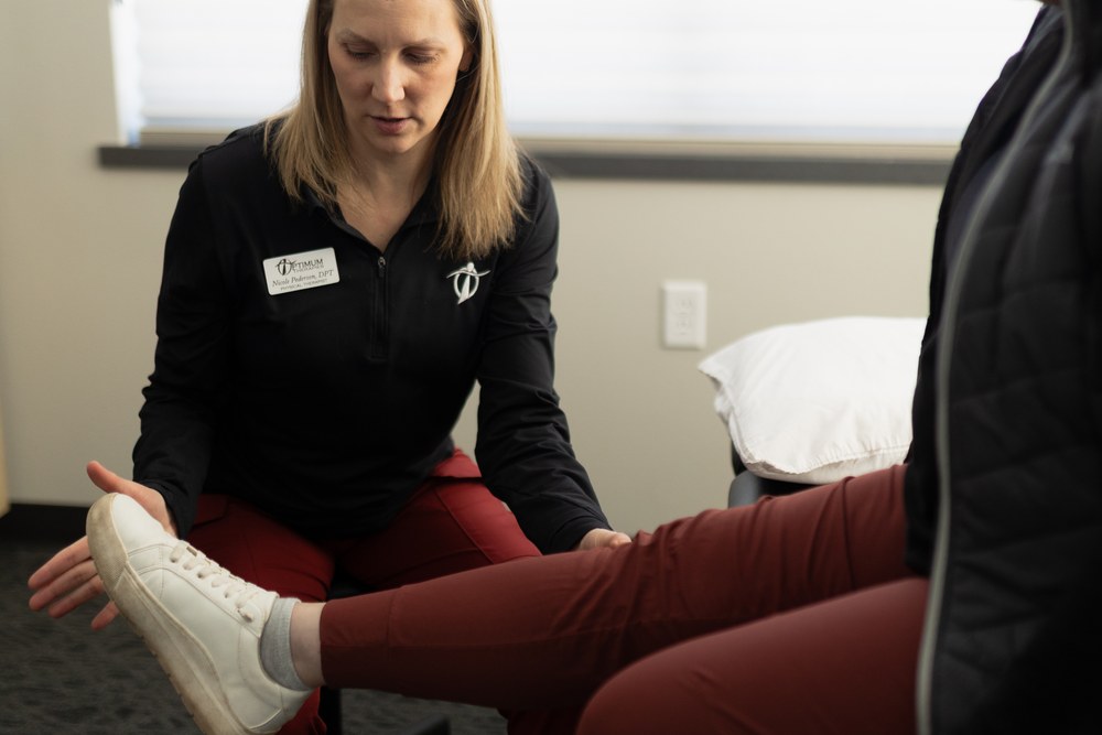 A physical therapist examines a patients leg, holding the ankle and knee while the patient sits on an exam table. The therapist wears a name tag and red pants, and both are indoors in a well-lit room.