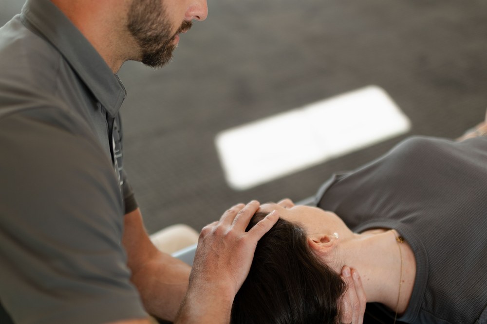A person lying on their back receives a neck adjustment from another individual, who gently supports their head and neck with both hands. Both are wearing gray shirts. The background is out of focus.