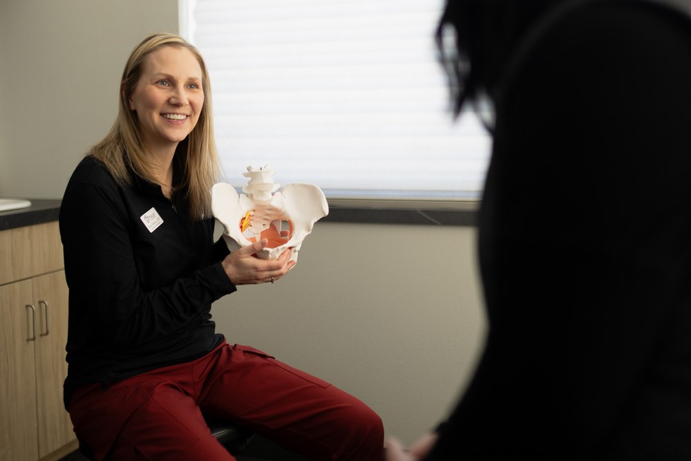 A smiling woman in medical attire holds a pelvic model while talking to another person in an examination room. The focus is on the woman explaining, with light coming from a window behind her.