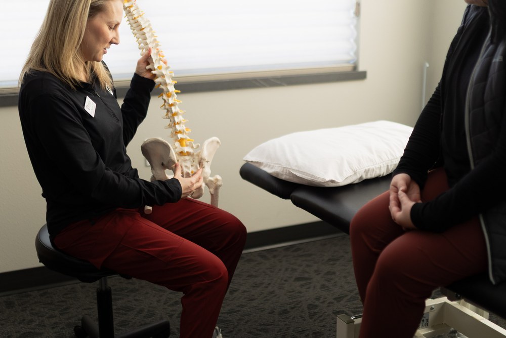 A healthcare professional holds a model of a human spine while explaining something to a seated patient in a medical office. A pillow rests on an examination table nearby.