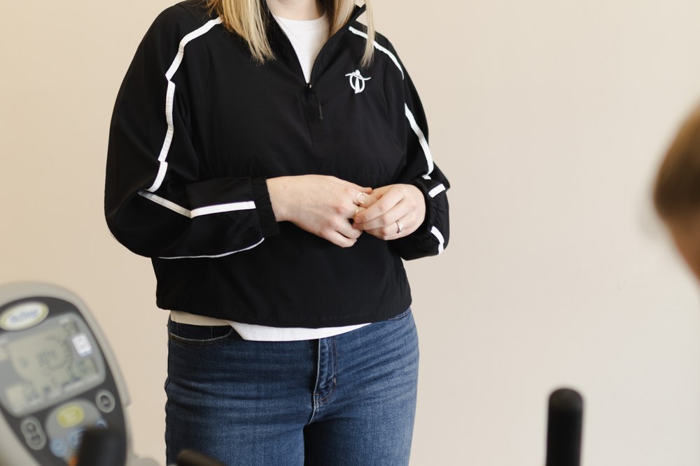 A woman wearing a black jacket with white stripes and blue jeans stands indoors. Her face is partially out of frame. Exercise equipment is visible in the foreground.