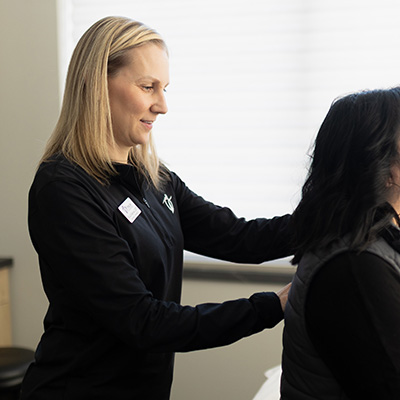 A woman with blonde hair, wearing a black top, examines or assists another seated woman with dark hair, who is facing away, in a brightly lit room.