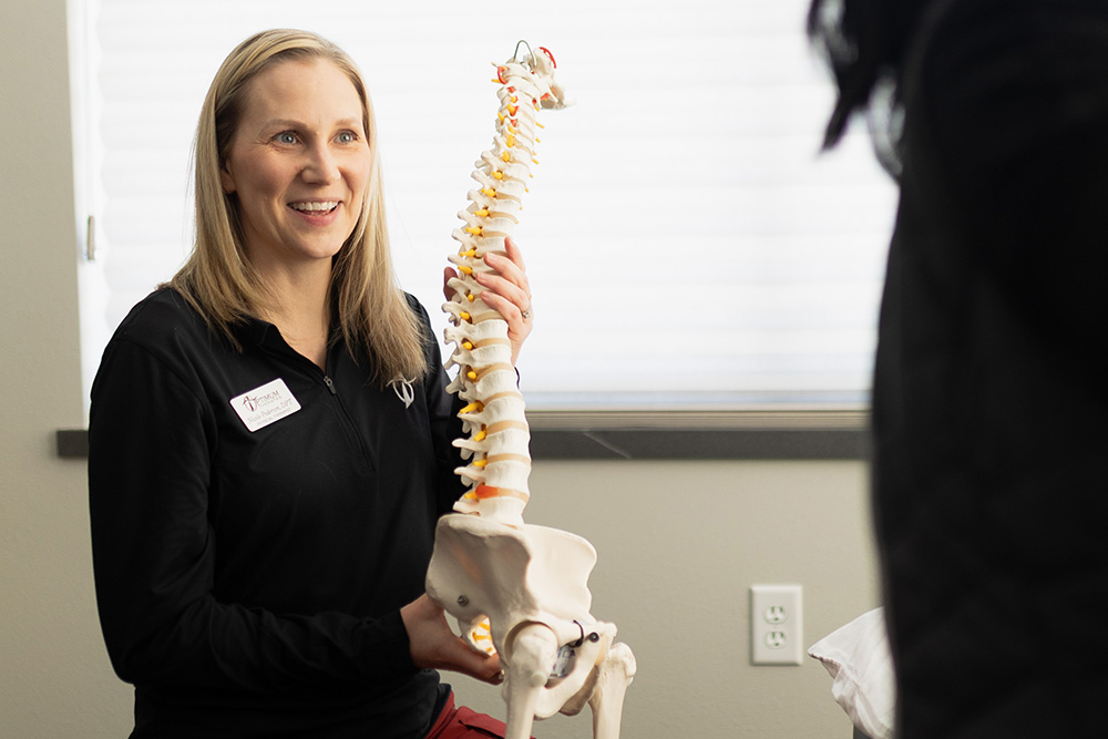 A woman smiles while holding a model of a human spine and pelvis, explaining something to another person in a well-lit room.