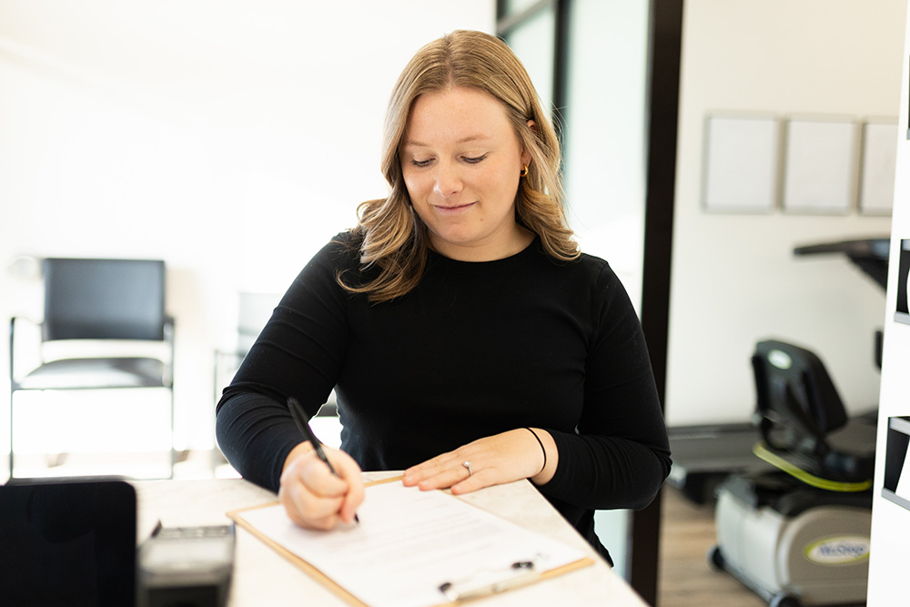 A woman with light brown hair in a black shirt is filling out a form on a clipboard at a counter in a bright, modern office or clinic setting.