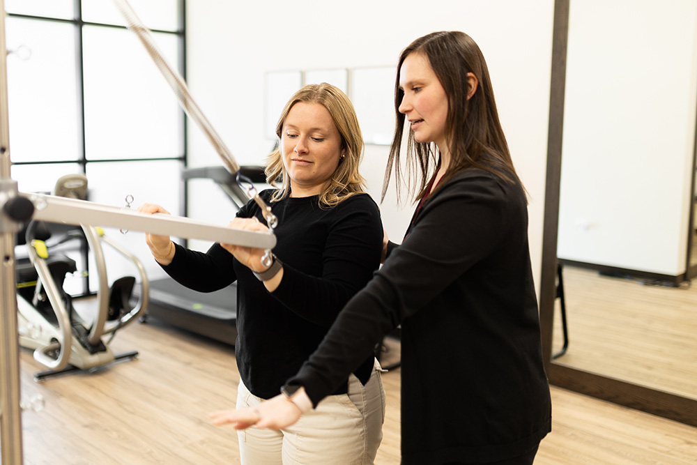 Two women in a gym, one guiding the other on how to use a fitness machine. Both are focused, standing on a wooden floor with exercise equipment in the background.