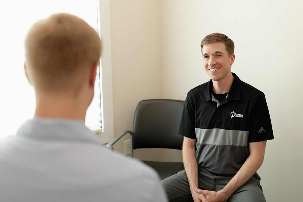 A man in a black and gray polo shirt sits and smiles while talking to another person in a bright, minimally furnished room.