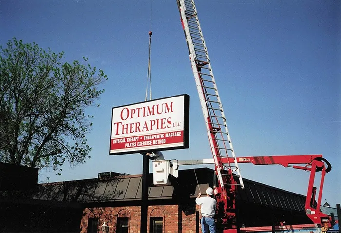 A worker on a cherry picker installs a large sign reading Optimum Therapies LLC outside a brick building under a clear blue sky. A tall ladder and tree branches are visible nearby.