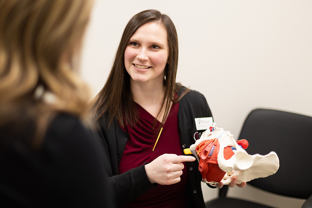 A woman in professional attire smiles while holding and pointing to a model of a pelvis during a discussion with another person in an office setting.