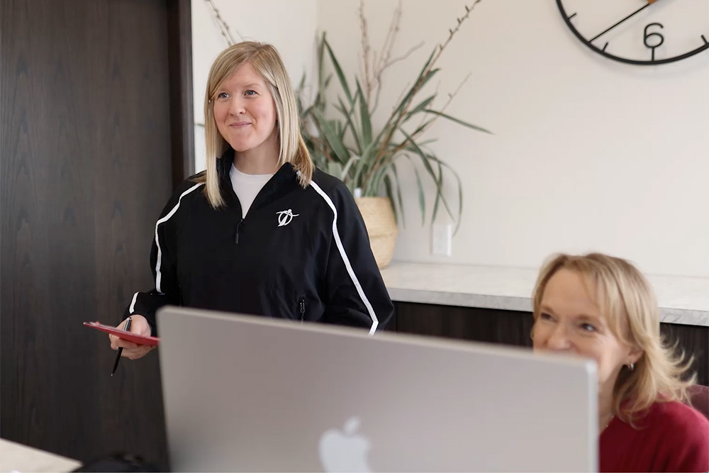Two women are in an office; one stands holding a tablet and smiling, while the other, partially visible behind a computer monitor, is seated and also smiling. Theres a plant and a clock in the background.