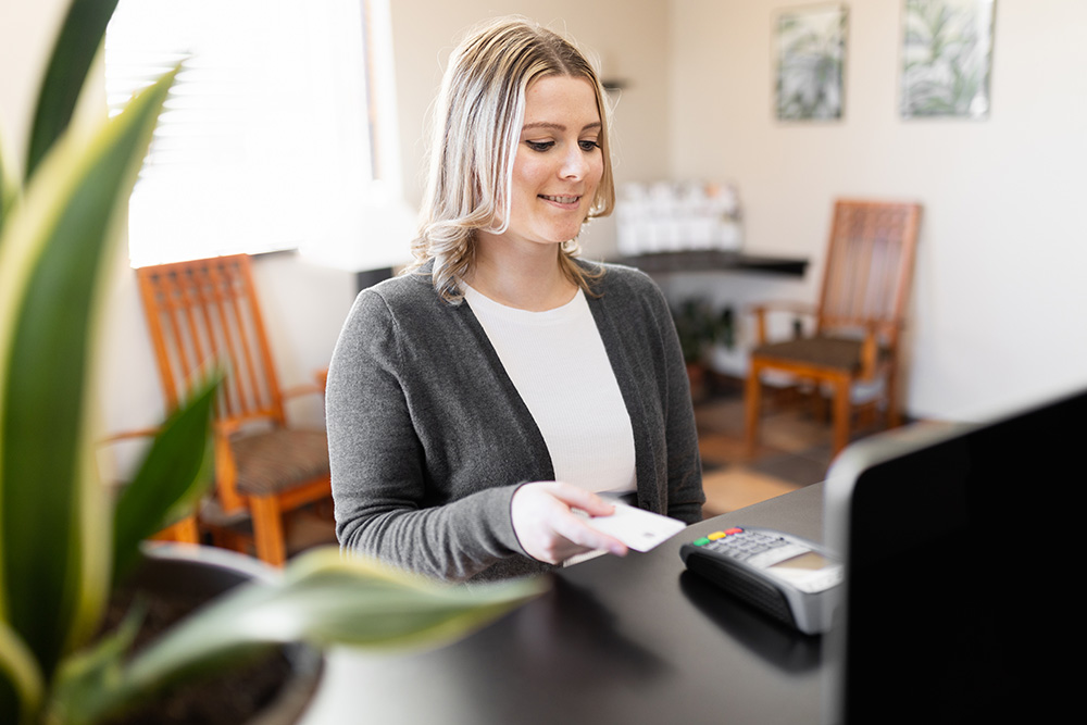 A woman sits at a desk, smiling while holding a card over a payment terminal. Wooden chairs and plants are visible in the bright, softly lit background.