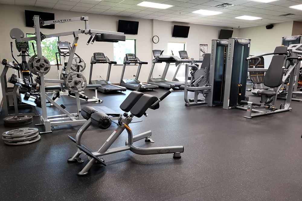 A gym with weight machines, free weights, treadmills, and other exercise equipment, in a clean, well-lit room with windows, TV screens, and a clock on the wall.