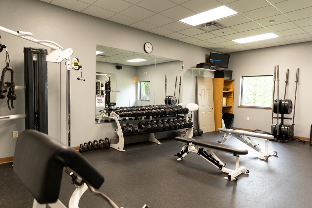 A gym with benches, dumbbells, weight racks, a mirror, and various exercise machines. There is a wall clock above the mirror and a window letting in natural light.