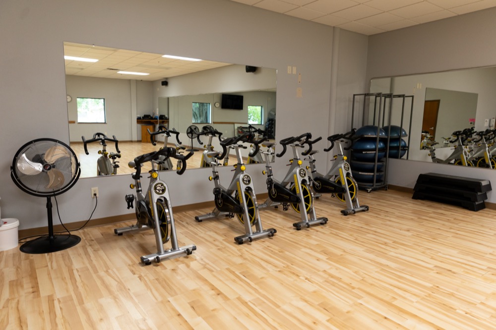 A fitness studio featuring a row of four indoor cycling bikes on a light wood floor. A black pedestal fan stands to the left, while a rack of stability balls and a step platform are located against the back wall, reflected in a large mirror.