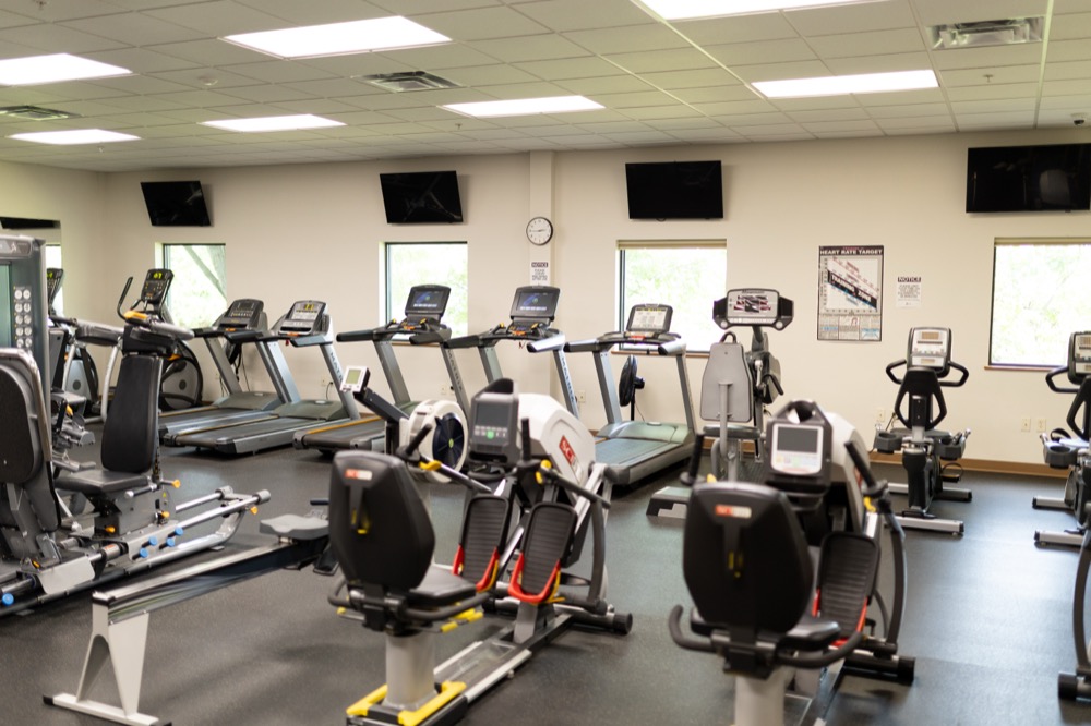 A gym with several treadmills, stationary bikes, and elliptical machines arranged neatly. The room has large windows, wall-mounted TVs, and exercise posters on the walls. The floor is covered with black rubber matting.