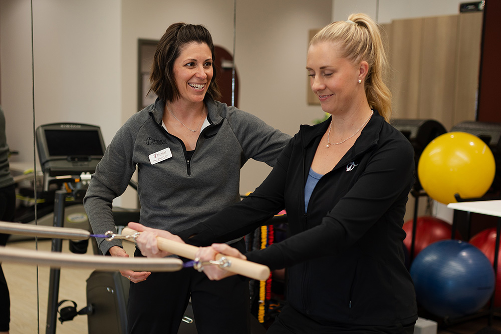 Two women in athletic wear are in a gym. One woman is assisting the other with a resistance band exercise. Colorful exercise balls and gym equipment are visible in the background.